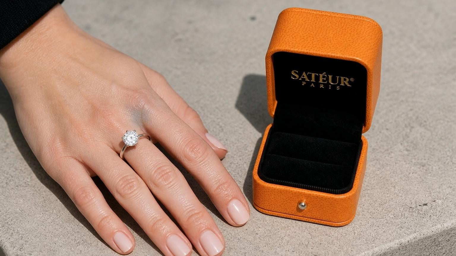 Engagement ring on fourth finger left hand with open Satéur ring box on concrete