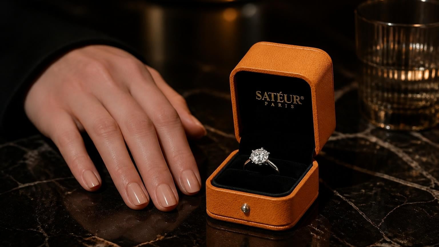 A Satéur Gems diamond simulant ring beside an open orange Satéur ring box on dark marble